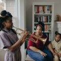 A child plays the trumpet indoors while family observes, creating a warm, musical atmosphere.
