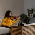 A girl in yellow sweater practicing violin in a warm home setting.