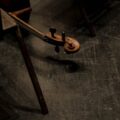 Artistic close-up of a cello scroll on a shadowed wooden floor with chairs nearby.