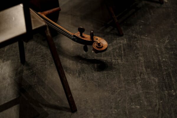 Artistic close-up of a cello scroll on a shadowed wooden floor with chairs nearby.