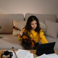 Young girl in yellow sweater learning violin using a tablet on a cozy sofa.