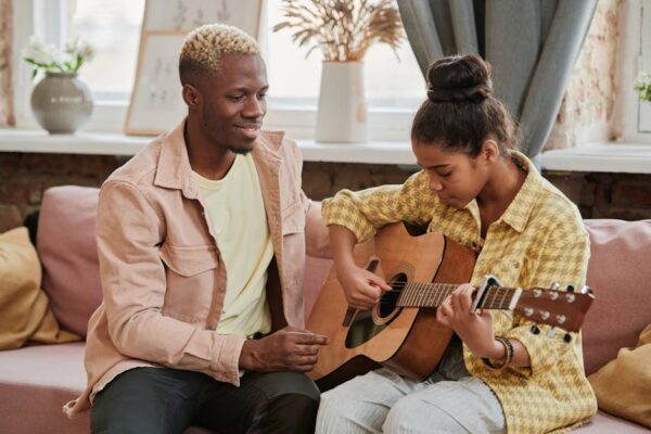 A father teaching his daughter to play acoustic guitar on a cozy indoor couch.