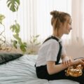 A young girl sitting on bed indoors, holding a saxophone, surrounded by plants.