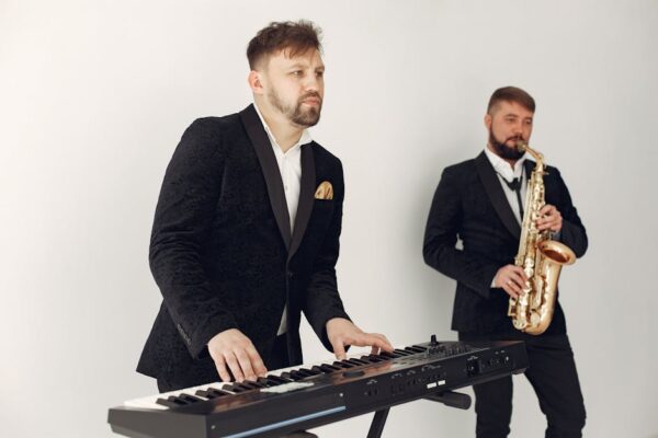 Two male musicians in suits playing a keyboard and saxophone in a studio setting.