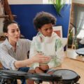 Teacher guiding a child in playing electronic drums with headphones indoors.