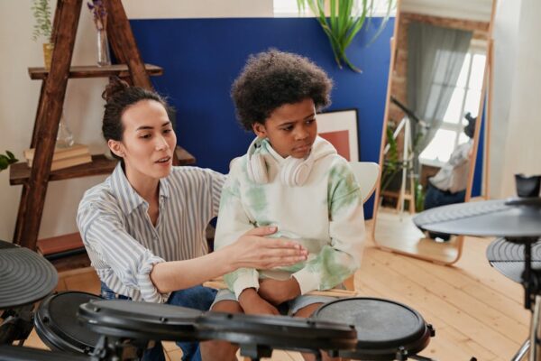 Teacher guiding a child in playing electronic drums with headphones indoors.