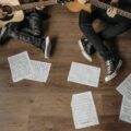 Two musicians playing guitars surrounded by sheet music on a wooden floor.