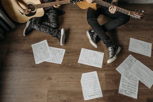 Two musicians playing guitars surrounded by sheet music on a wooden floor.