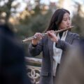 Asian woman in a blazer playing flute outdoors with a blurred background.