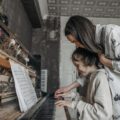 A mother guides her daughter in learning piano amidst a vintage setting, creating a warm educational moment.