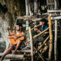 Young ethnic male guitarist playing classic guitar near cheerful partner with djembe on bamboo staircase near tree and looking away