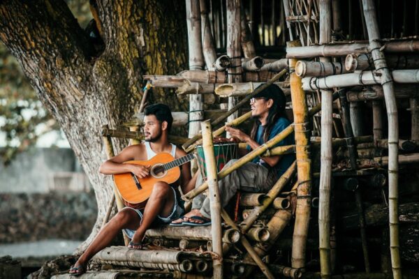 Young ethnic male guitarist playing classic guitar near cheerful partner with djembe on bamboo staircase near tree and looking away