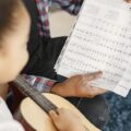 A young girl learning guitar with a teacher, focusing on a music sheet.