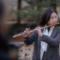 A woman in a business suit plays the flute in an outdoor park setting, showcasing musical talent.