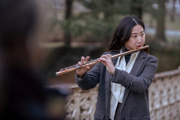 A woman in a business suit plays the flute in an outdoor park setting, showcasing musical talent.