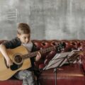 A young boy practicing guitar indoors on a comfy leather sofa, focusing on his music sheet.