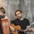 A young boy learning guitar from a bearded man indoors, emphasizing musical guidance.