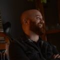Smiling bearded man in casual wear, standing by guitar indoors.
