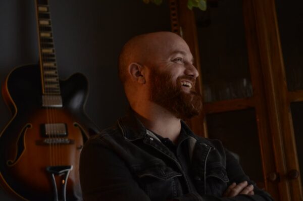 Smiling bearded man in casual wear, standing by guitar indoors.