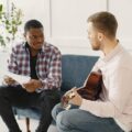 Two men sitting on a couch discussing music with guitar and papers.