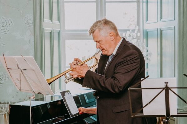 Side view of concentrated senior musician with grey hair in dark elegant suit looking at notes on sheet and playing trumpet next to pianist