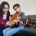 A father tutoring his son on acoustic guitar playing at home, fostering a musical bond.