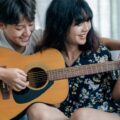 A joyful lesbian couple enjoys playing an acoustic guitar together indoors, showcasing their close relationship and shared passion for music.