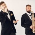 Two male musicians in black suits performing with a saxophone and microphone on a studio background.