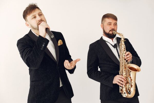 Two male musicians in black suits performing with a saxophone and microphone on a studio background.