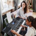 Girl takes piano lessons with attentive teacher observing indoors.