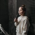 A young girl sings into a microphone in a soundproof room with acoustic panels around her.