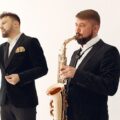 Talented male musician playing saxophone near inspired male vocalist singing beautiful song while standing together against white wall during celebration party or live music show