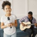 Father playing guitar as daughter sings joyfully with a microphone indoors.