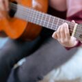 Soft focus of crop anonymous female musician in casual outfit playing acoustic guitar while sitting on pouf