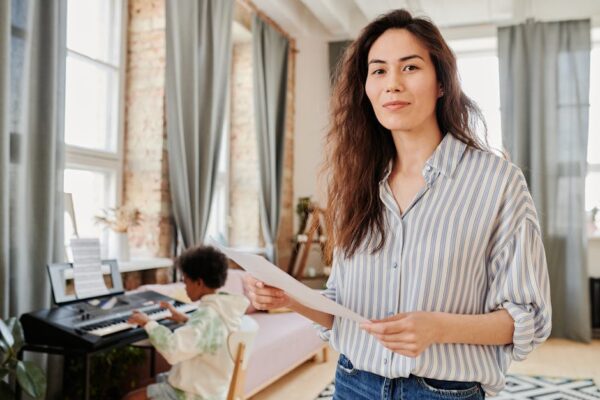 A music teacher assists a child learning piano indoors in a bright room.