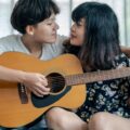 A loving couple shares a tender moment while playing guitar together indoors.