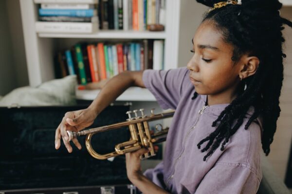 A focused young musician plays the trumpet indoors near a bookshelf.