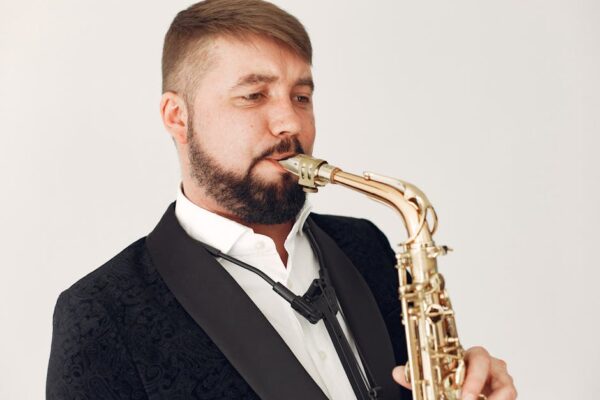A bearded musician plays the saxophone, dressed in an elegant black suit and white shirt.