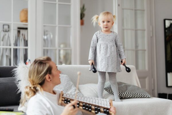Young girl standing on couch holding percussion instruments, enjoying playful music time with adult nearby.