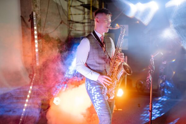 Saxophonist playing in a colorful, dynamic concert with smoke and lights.