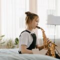 A young girl in overalls playing the saxophone indoors, focusing on music practice.