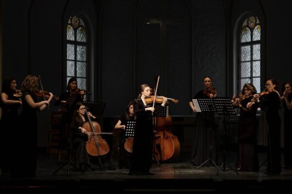 Women musicians perform with violins and cellos in a classical orchestra setting.