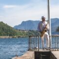 A man playing the saxophone on a pier by a lake, surrounded by mountains on a sunny day.