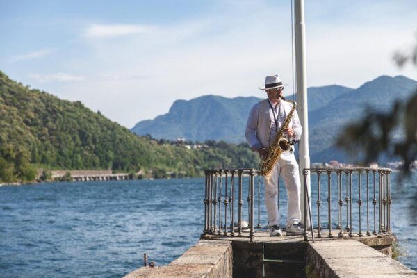 A man playing the saxophone on a pier by a lake, surrounded by mountains on a sunny day.