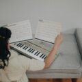 A young girl sits indoors playing an electronic keyboard with music sheets.