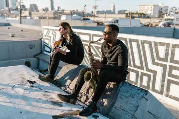 Two musicians playing guitar and saxophone on a sunny rooftop with urban landscape.