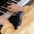 From above of crop unrecognizable female musician in warm pullover sitting on piano stool and touching black and white keys