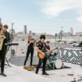 A diverse music band plays on a rooftop with city skyline in the background.
