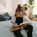 A young girl sits on a bed writing music notes while holding a saxophone.