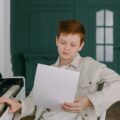 A young child reading sheet music while sitting at a piano indoors, focusing on practice.